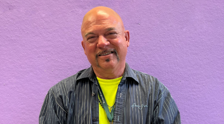 A picture of David smiling. He is wearing a yellow t-shirt with a striped navy blue button up on top. 